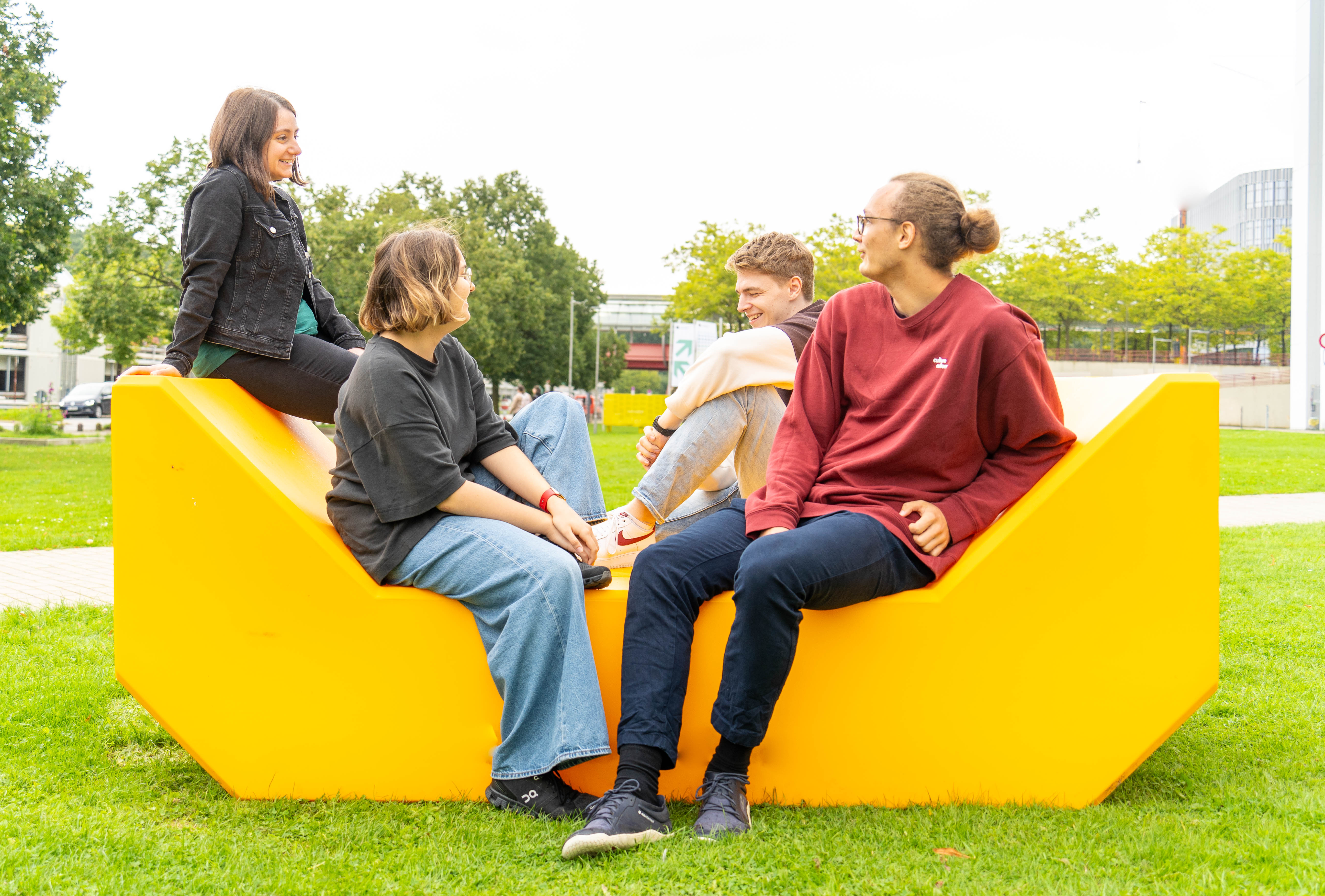 Vier Studierende sitzen auf einer gelben Bank auf der Wiese vor der Uni